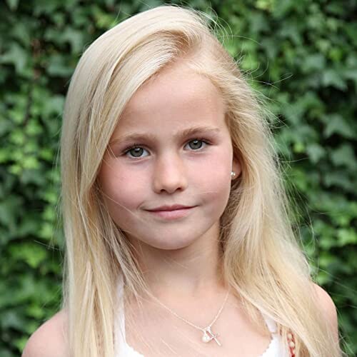 Young girl with blonde hair standing outdoors in front of greenery.